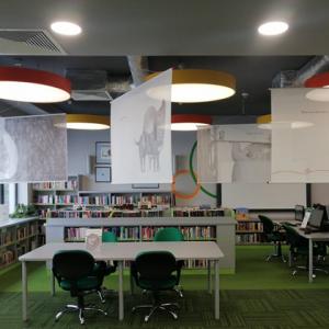 Widok na salę biblioteczną, pod sufitem zawieszone plansze wystawy z ilustracjami z książki "Co robią uczucia?".