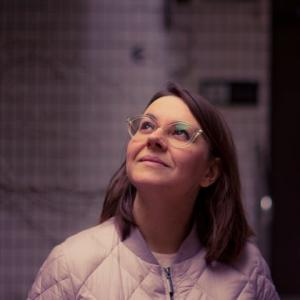 Fotografia Aleksandry Cieślak. Uśmiechnięta kobieta spogląda w górę, jest ubrana w kurtkę na nosie ma okulary.