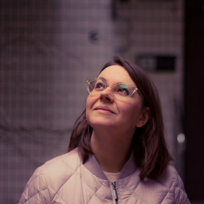 Fotografia Aleksandry Cieślak. Uśmiechnięta kobieta spogląda w górę, jest ubrana w kurtkę na nosie ma okulary.