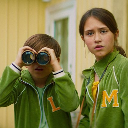 Kadr z filmu: dwójka przyjaciół dziewczynka i chłopiec patrzą przed siebie, chłopiec spogląda przez lornetkę. Na sobie mają zielone bluzy, ona z dużą literą „M”, on z „L”. W tle widać dom.