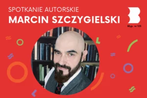 Uśmiechnięty, ogolony na łyso, brodaty mężczyzna patrzy śmiało na lewo w obiektyw, na tle półek pełnych książek.
