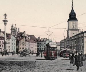 Na fotografii widoczny jest tramwaj na środku placu, a w tle konie zaprzężone do powozów oraz przechodnie. Obok znajdują się historyczne budynki, w tym jeden z charakterystyczną, wysoką wieżą. Centralnym punktem placu jest również monument z posągiem na kolumnie. Całość tworzy obraz tętniącego życiem, historycznego centrum miasta.