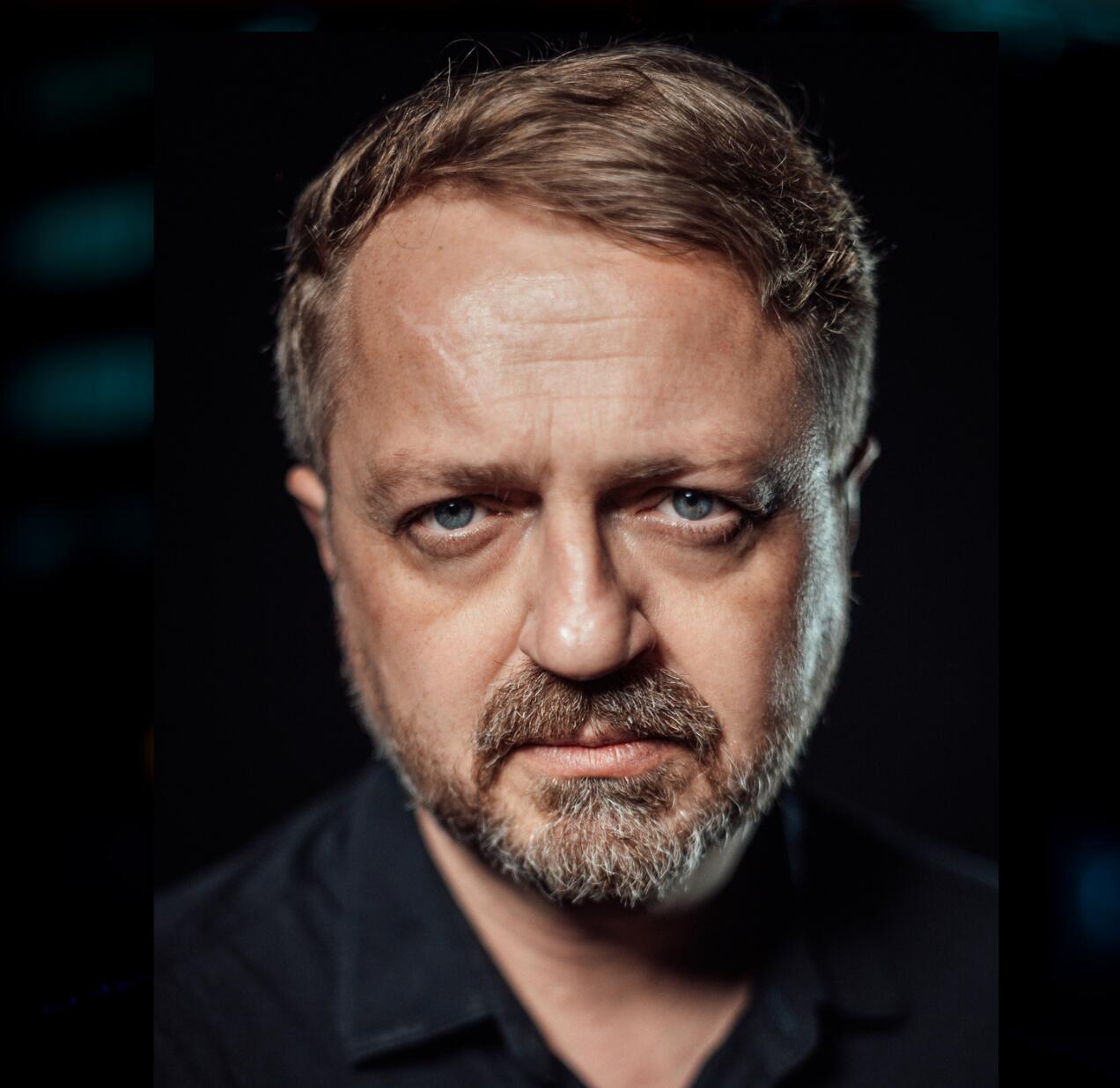 Przedstawiam portret Przemysława Borkowskiego. Jest to zbliżenie na twarz dojrzałego mężczyzny. Jego oczy są intensywnie niebieskie, a na twarzy widoczny jest zarost. Włosy są krótkie i w kolorze brązowym. Ubrany jest w ciemną koszulę. Patrzy prosto w obiektyw z poważnym wyrazem twarzy.