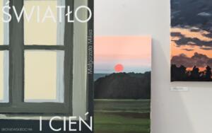 Na załączonym obrazku widać fragment wystawy sztuki, prezentującej kilka obrazów. Po lewej stronie znajduje się duży obraz lub plakat, który przedstawia stylizowaną ramę okienną w odcieniach szarości i beżu. Na nim widnieje duży biały napis "ŚWIATŁO" u góry oraz "I CIEŃ" na dole. Pionowo, po prawej stronie tego obrazu, jest napis "Małgorzata Miksa". W lewym dolnym rogu widoczny jest również adres "BRONIEWSKIEGO 9A". Obok, w centralnej części kadru, wisi mniejszy obraz przedstawiający pejzaż. Ukazuje on zachód lub wschód słońca nad horyzontem, z wyraźnym, czerwonym słońcem na tle różowo-pomarańczowego nieba, przechodzącego w błękit. Poniżej widać ciemne sylwetki wzgórz lub lasu oraz zielone pole na pierwszym planie.